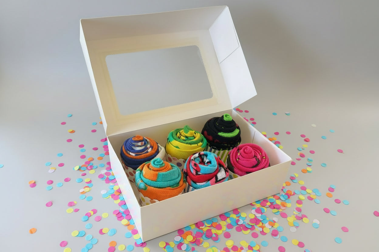 Box of colorful sock cupcakes with a window on top, surrounded by confetti on a light background