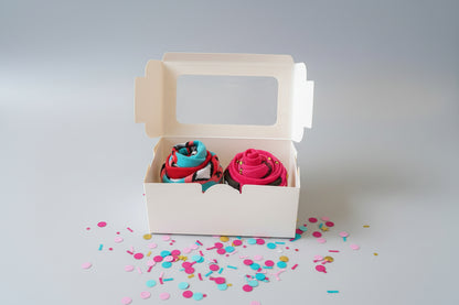 Two colorful sock cupcakes in a small white box with confetti on a light gray background